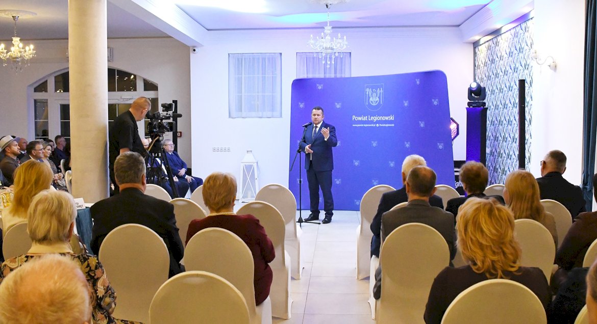 Zdjęcie - na pierwszym planie tyłem zgromadzeni goście wydarzenia słuchający prelekcji, w tle starosta legionowski przy mikrofonie na tle ścianki promocyjnej Powiatu Legionowskiego.