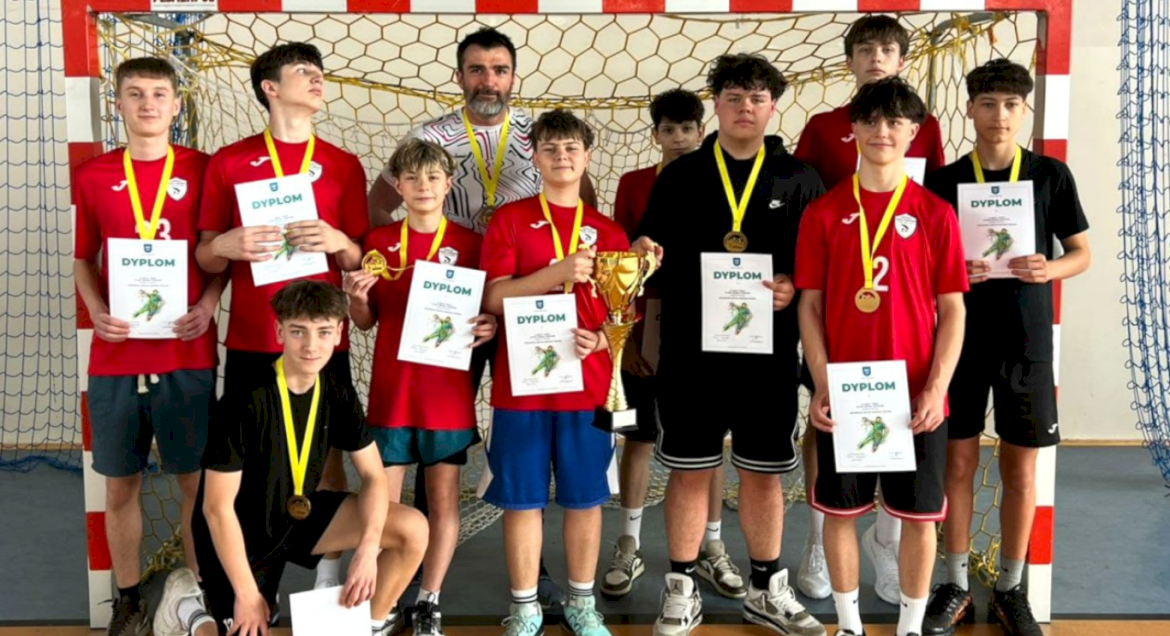 Na zdjęciu w bramce stoi cała drużyna chłopców z medalami na szyjach, a pośród nich stoi też mężczyzna z czarną brodą, czyli ich trener