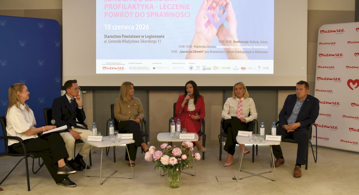 „Panel dyskusyjny poświęcony onkologii z udziałem sześciu prelegentów siedzących na scenie m.in. członkini Zarządu Województwa Mazowieckiego Anna Brzezińska, starosta legionowski Roman Smogorzewski oraz wójt gminy Nieporęt Agnieszka Powała.W tle wyświetlona jest plansza informująca o konferencji „Droga do zdrowia – profilaktyka, leczenie, powrót do sprawności”, która odbędzie się 18 czerwca 2026 roku w Legionowie.”
