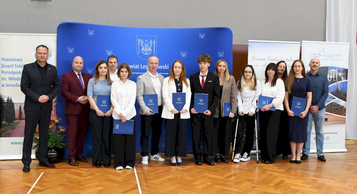 na zdjęciu grupa osób podczas rozdania nagród starosty - młodzież, rodzice oraz przedstawiciele władz Powiatu na tle powiatowej ścianki