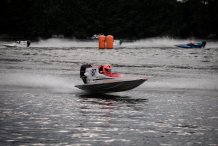 Zdjęcie przedstawia sportową motorówkę z numerem 87 mknącą po tafli jeziora. Sternik ubrany w czerwony kask. Zdjęcie wykonane podczas zawodów. Na wodzie rozieszczone elementy wyznaczające trasę.