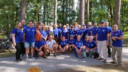 Na chodniku stoi grupa kilkunastu osób, kobiet i mężczysn, uczestników rajdu. Większośc ma okolicznościowe koszulki w kolorze granatowym. W tle las.