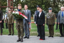 Zdjęcie: na pierwszym planie w centralnym punkcie stoi żołnierz trzymający wieniec z biało-czerwoną szargą, na drugim planie stoją przedstawiciele wojska, władz samorzadowych i inni zaproszeni goście.