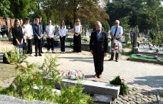 Zdjęcie wykonane na cmentarzu w Legionowie. Na pierwszym planie mężczyzna oddający hołd pod pomnikiem  „Oddali życie na nieludzkiej ziemi za Ojczyznę i swoje miasto”. Na drugim plenie grupa jedenastu osób, głównie młodzieży licealnej. W tle nagrobki.