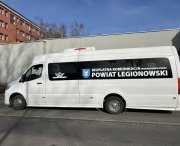 Zdjecie przedstawia biały autobus, stojący na przystanku,  z oznakowaniem Bezpłatnych Powiatowych Linii Autobusowych