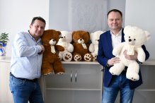 na zdjęciu dwie osoby, w ręcach mają duże, pluszowe misie, które też sa poustawiane na szafce z tyłu