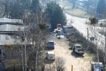 Widok z góry - po lewej stronie: trzy budynki do rozbiórki; po prawej stronie nieutwardzona droga oraz samochody dostawcze. Po obu stronach zieleń.
