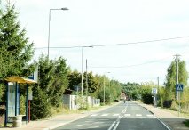 Zdjęcie: fragment drogi powiatowej nr 1810W w Gminie NIeporet, widok na asfalt, przejście dla pieszych oraz przystanek autobusowy. Po obu stronach roślinność oraz zabudowania.