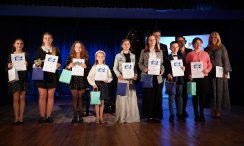 Na zdjęciu znajduje się grupa dzieci i młodzieży stojąca na scenie, trzymająca dyplomy i torby. W tle widoczny jest fortepian oraz troje dorosłych organizatorów i nauczycieli..