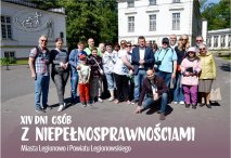 Na zdjęciu grupa uczestników XIV Dni Osób z Niepełnosprawnościami Miasta Legionowo i Powiatu Legionowskiego. Uczestnicy stoją wspólnie przed zabytkowym budynkiem kompleksu Pałacowo-Parkowego w Jabłonnie, w słoneczny dzień,