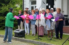 Na zdjęciu grupa ludzi śpiewa, przed nimi mikrofony, za nimi budynek MOK
