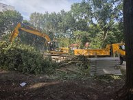 Zdjęcie przedstawia koparkę podczas prac rozbiórkowych na zadrzewionym terenie.