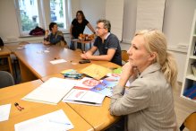 Zdjęcie: grupa osób pracuje przy stolikach, na których leżą materiały.