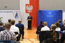 Zdjęcie wykonane na sali gimnastycznej LO im. Konopnickiej w Legionowie. Na pierwszym planie, tylem, zebrana publicznośc, w tle przy mównicy mężczyzna, za nim flaga państwowa oraz ścianki promocyjne.