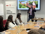 Uczestnicy szkolenia „Szkoła Turystyki Zrównoważonej” siedzą przy stołach konferencyjnych. Na pierwszym planie widoczna jest kobieta notująca w zeszycie, w tle trener omawia zagadnienia, korzystając z rzutnika.