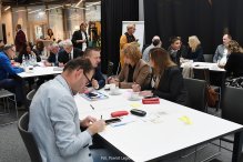 Na zdjęciu osoby siedzące przy stoliku. W tle widać więcej osób