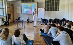 Grupa uczniów siedzi na sali. Na drugim planie osoba prowadzaca zajęcia.