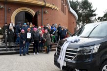 Zdjęcie: na pierwszym planie maska mikrobusu z kokardą na dugim planie sotjąca na schodach grupa osób, w tle kościół.