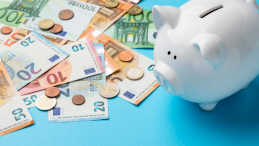 Na zdjęciu widać na niebieskim tle rozłożone pieniądze i białą świnkę skarbonkę