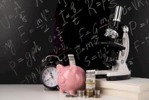 Na zdjęciu czarna tablica kredowa, a przed nią skarbonka świnka, zwinięty rulon pieniędzy, zegarek i mokroskop