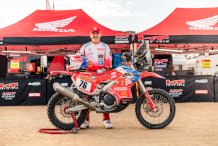 Na zdjęciu widoczny jest biało-czerwony motocykl i jego kierowca, który stoi za nim