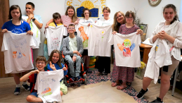 Na zdjęciu grupowym znajdują się podopieczni Fundacji Promień Słońca, wszyscy trzymają w rękach białe koszulki z kolorowymi rysunkami wykonanymi własnoręcznie