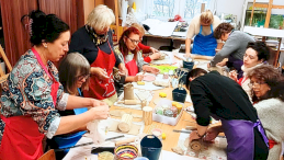 Na zdjęciu widać jak uczestniczki warsztatów ubrane w fartuchy siedzą a część stoi przy stole, na którym znajduje się glina, farby i wiele przyrządów do wykonywania talerzy ceramicznych