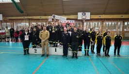 Powiększ obraz:Dziewczęta w czarnych uniformach strażackich stojące na trzech stopniach podium oznaczonych numerami od 1 do 3. Każdy ze stopni podium to miejsce w zawodach i na każdym z nich stoi kilka - kilkanaście dziewczynek. Przed nimi dorośli strażacy reprezentujący organizatorów zawodów.