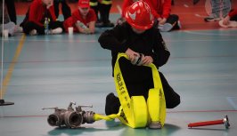 Powiększ obraz:Dziecko w hełmie strażackim klęczące na podłodze w hali sportowej, zajmujące się wężem strażackim.