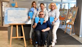 Powiększ obraz:Grupa pracowników starostwa, którzy mają założone niebieskie elementy garderoby, stoją w holu, jedna osoba na wózku, w tle niebieskie serduszka i tablica