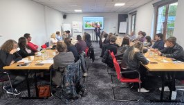 Powiększ obraz: Sala szkoleniowa podczas zajęć „Szkoły Turystyki Zrównoważonej”. Uczestnicy siedzą przy długich stołach i słuchają trenera, który prowadzi prezentację wyświetlaną na ekranie. Na ekranie widoczny jest obraz średniowiecznych murów obronnych. Po lewej stronie ustawiony jest roll-up z logotypem Ministerstwa Sportu i Turystyki i nazwą projektu.