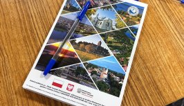 Powiększ obraz: Notes z logo projektu „Szkoła Turystyki Zrównoważonej” leży na drewnianym stole. Na okładce znajdują się kolorowe fotografie przedstawiające krajobrazy i zabytki regionu Jeziora Zegrzyńskiego, ułożone w trójkątnych ramkach. Na dole widoczne są logotypy Ministerstwa Sportu i Turystyki oraz informacja o dofinansowaniu zadania. Na notesie położony jest niebieski długopis.