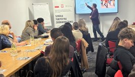 Powiększ obraz: Grupa uczestników szkolenia „Szkoła Turystyki Zrównoważonej” siedzi przy stołach konferencyjnych i słucha wykładu. Trener stojący przy ekranie wskazuje schemat dotyczący turystyki zrównoważonej, obok którego widoczna jest fotografia tańczącej grupy ludzi. W tle roll-up z logo Ministerstwa Sportu i Turystyki oraz tytułem projektu.